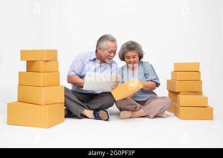 Asiatique senior couple tenant boîte de colis et ordinateur portable et assis sur le sol isolé sur fond blanc, en ligne marketing boîte de livraison co Banque D'Images