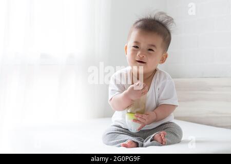 Bébé asiatique tenant le lait en bouteille et assis sur un lit blanc Banque D'Images