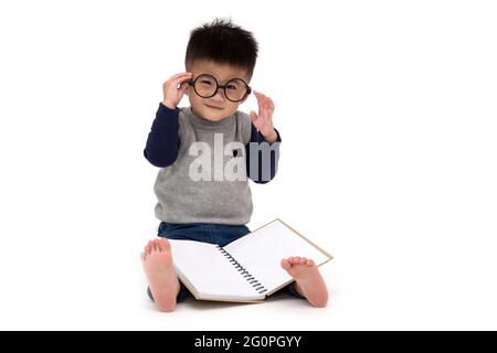 Portrait d'un adorable petit garçon asiatique portant des lunettes assis sur le sol et lisant un livre isolé sur fond blanc, concept de rentrée scolaire, 1 an Banque D'Images