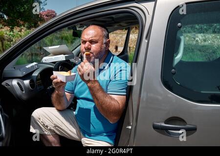 Drôle barbu homme d'âge moyen manger un sandwich vegan dans la rue près de la voiture. Manger malsain. Restauration rapide. Plats à emporter Banque D'Images