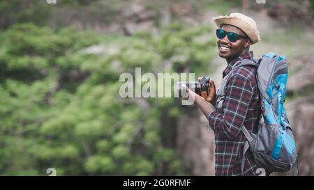 Homme africain voyageur tenant un appareil photo avec sac à dos debout sur le sommet de la montagne Banque D'Images