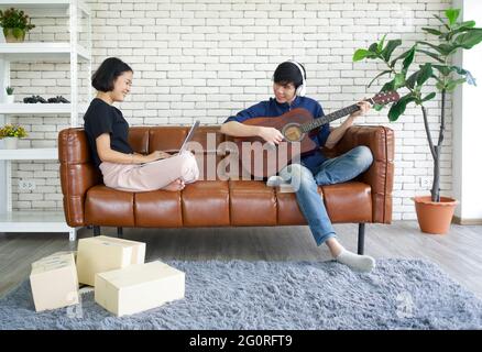 Les jeunes amoureux passent du temps ensemble en vacances dans le salon. Homme asiatique avec casque jouant une guitare acoustique pendant que la fille vérifie la parcelle Banque D'Images