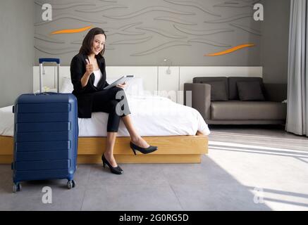 Femme d'affaires asiatique en costume noir assise avec un ordinateur tablette dans la main. Ses bagages ont été placés à côté du lit. Atmosphère de travail à l'extérieur pendant le Banque D'Images