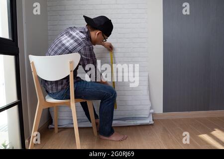 Jeune homme asiatique encollant des papiers peints sur le mur à la maison. Salle de rénovation dans la maison par auto-bricolage concept Banque D'Images