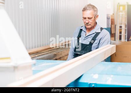 Portrait d'un menuisier âgé ou d'un charpentier en combinaison travaillant avec des planches en bois dans un atelier de menuisier. Sciage de bois. Banque D'Images