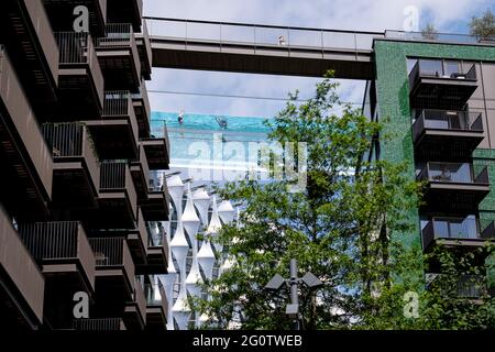 Avec l'architecture de l'ambassade américaine ci-dessous, les nageurs apprécient les eaux de la piscine Sky, une piscine d'eau transparente de 25 mètres de long reliant deux tours résidentielles de 10 étages à 35 mètres au-dessus du sol, La plus grande structure de piscine en acrylique autonome au monde dans le nouveau développement Embassy Gardens d’EcoWorld Ballymore à neuf Elms, le 3 juin 2021, à Londres, en Angleterre. Banque D'Images