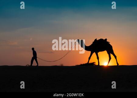 Désert de Thar, Rajasthan, Inde - octobre 15th 2019 : Silhouette d'un caméléer menant un chameau dans des dunes de sable. Coucher de soleil avec ciel bleu en arrière-plan Banque D'Images