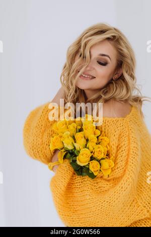 Portrait d'une belle jeune femme blonde souriante avec un bouquet de roses jaunes dans un chandail volumineux sur fond blanc dans le studio. Soft se Banque D'Images