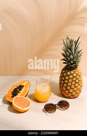 un verre rafraîchissant de jus de fruit avec une paille et un ananas, une demi-papaye, un demi-pamplemousse et une paire de grandes lunettes de soleil sur le sable o Banque D'Images