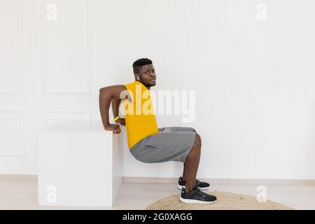 Vue sur toute la longueur jeune élégant coupe forte Afro américain homme dans des vêtements de sport jaunes profondément squatting à l'intérieur à la maison ou en studio. Homme millénaire en bonne santé Banque D'Images