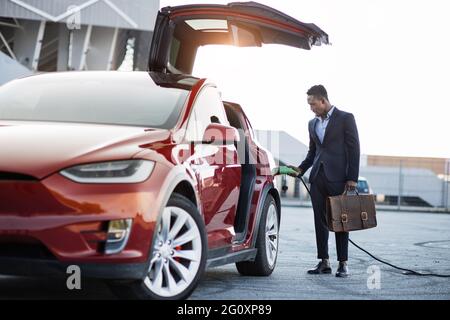 Portrait complet d'un bel homme africain en costume d'affaires tenant une valise et chargeant une voiture électrique à l'extérieur. Concept de personnes, modes de vie modernes et véhicule écologique. Banque D'Images