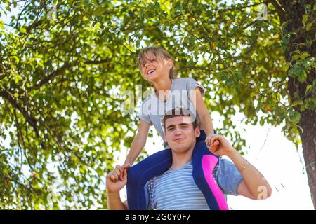 Frère défoqué de la sœur de circonscription à l'arrière. Portrait d'une fille heureuse sur les épaules de l'homme, porcgyback. Petite mouche, levez la main. Famille jouant à l'extérieur. Arbre vert Banque D'Images