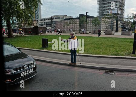 Londres (Royaume-Uni): Un homme avec une béquille marche entre les véhicules attendant aux feux de signalisation à Elephant et Château mendiant. Banque D'Images