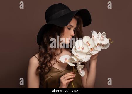 Belle femme aux cheveux longs et sains bruns bouclés. Magnifique modèle avec maquillage naturel et coiffure parfaite portant un chapeau classique tenant des fleurs blanches Banque D'Images