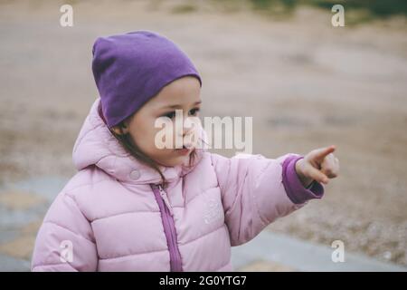 Petite fille 3-4 dans la veste rose et chapeau lilas points quelque part sur une route de terre dans la campagne Banque D'Images