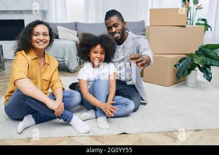 La famille afro-américaine s'installe dans une nouvelle maison. Le père, la mère et la fille sont heureux de s'asseoir sur un plancher de maison après avoir déballé les choses. Achat ou renti Banque D'Images