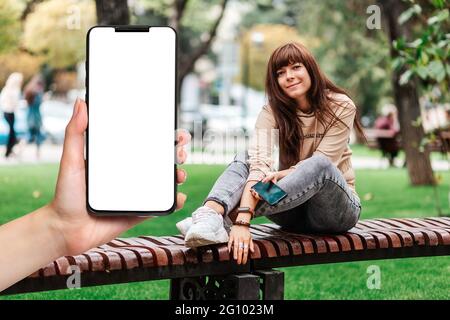 La main d'une femme tient un smartphone avec un écran blanc, gros plan. Une jeune femme est assise sur un banc et utilise un smartphone. Le concept de zone wi-fi gratuite. Banque D'Images