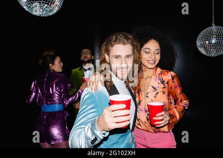 amis multiraciaux positifs dans des vêtements colorés avec des tasses en plastique dans les mains dansant sur fond noir Banque D'Images