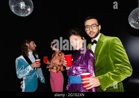 amis multiraciaux positifs dans des vêtements colorés debout avec des tasses en plastique dans les mains sur fond noir Banque D'Images