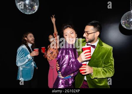 jeunes adultes amis multiraciaux dans des vêtements colorés avec des tasses en plastique dans les mains dansant sur fond noir Banque D'Images