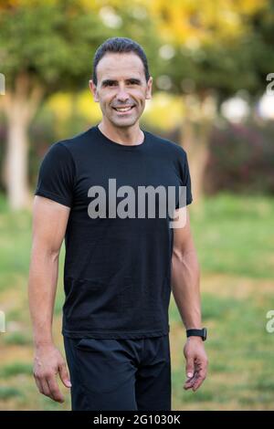 Homme adulte portant des vêtements de sport noirs dans le parc. Il sourit et regarde la caméra. Banque D'Images