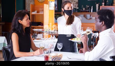 un bussboy féminin positif dans le masque servant l'ordre à l'homme et à la femme dans le restaurant moderne Banque D'Images