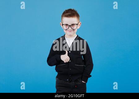 Comme. Portrait d'un jeune écolier heureux avec des lunettes souriant à l'appareil photo et faisant un geste de pouce vers le haut, montrant un signe d'approbation sympa d'accord. Studio d'intérieur isolé sur fond bleu. Banque D'Images