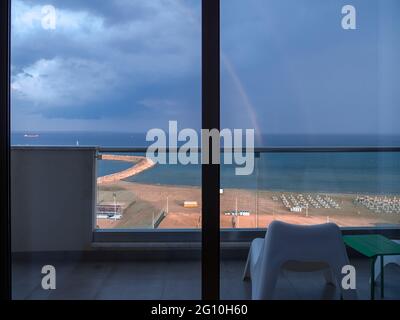 Vue aérienne supérieure surplombant l'arc-en-ciel au-dessus de la plage méditerranéenne avec chaises longues près de la promenade des palmiers de Finikoudes dans la ville de Larnaca. Ciel sombre et nuageux. Banque D'Images