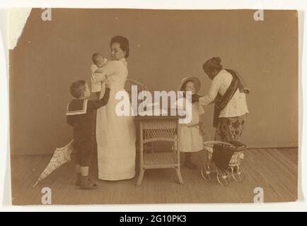 Trois enfants avec mère et leur basoe. À gauche d'une table et d'une chaise une femme debout en costume d'infirmière blanc avec un bébé sur le bras. En plus d'elle un garçon en costume de marin avec un parapluie indiquant une sucette. A droite une fille Mret une poupée pram qui obtient un chapeau mis en place par une femme indonésienne à Sarong et Kabaiah. Banque D'Images