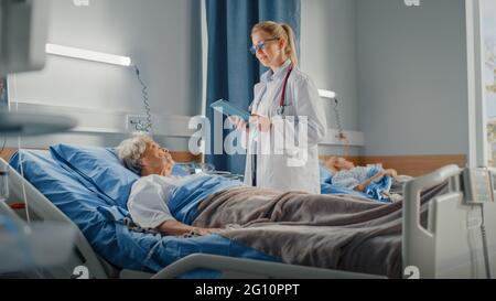 Service hospitalier : un médecin amical pose des questions de soins de santé à une dame âgée souriante qui se repose au lit. Le médecin utilise un Tablet PC, effectue une vérification, parle Banque D'Images