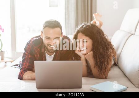 Un jeune couple se détend sur le canapé et magasine en ligne avec un ordinateur portable. Le couple engagé s'amuse avec l'ordinateur portable pour passer des appels vidéo Banque D'Images