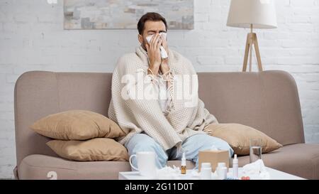 homme malade assis sur un canapé et éternuant dans un tissu près d'une table basse avec des boissons et des bouteilles Banque D'Images