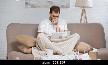 homme malade assis sur un canapé avec un ordinateur portable tout en toussant près d'une table basse avec des boissons et des bouteilles Banque D'Images