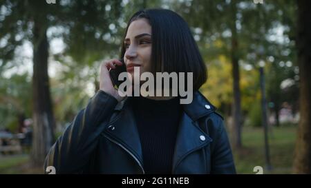 Parmi les arbres verts, la jeune femme qui marche dans le parc, parle bien au téléphone et sourit, aime une conversation avec son bien-aimé ou ami. Banque D'Images