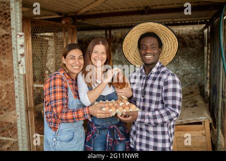 Ravi homme et femme multiracial avec du poulet et des œufs souriant et regardant la caméra tout en se tenant près de la houle de poule pendant le travail à la ferme Banque D'Images