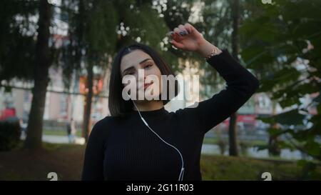 Souriante jeune belle femme avec dans le casque écoute de la musique et danse marchant le long du parc lent mouvement Banque D'Images