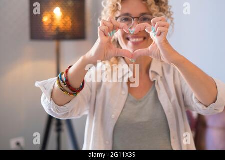 Joyeux portrait adulte beau de femme faisant la forme du coeur avec les mains devant l'appareil photo avec l'arrière-plan intérieur maison - gaie les gens de femme aiment Banque D'Images