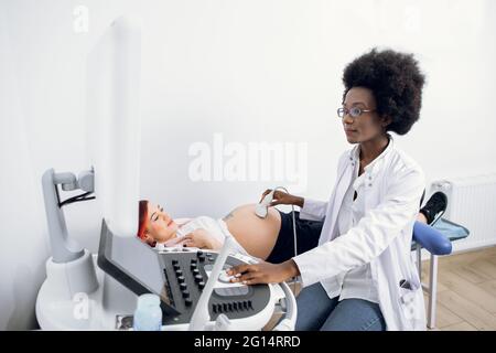 Jeune professionnel sympathique femme afro-américaine médecin faisant l'échographie 3d sur le ventre de la femme enceinte en clinique. Échographie pour femme enceinte. Banque D'Images