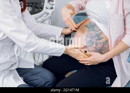 Image rognée en gros plan d'une femme médecin obstétricienne méconnue, examinant une jeune femme enceinte assise sur un canapé à l'hôpital. Mignon ventre enceinte avec fleurs tatouage. Palpation du ventre enceinte Banque D'Images