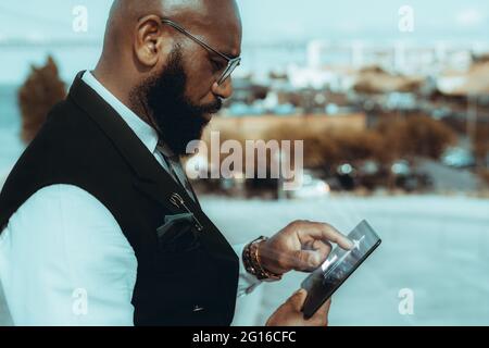 Une vue latérale d'un élégant homme d'affaires noir chauve et mûr entrepreneur en plein air dans des lunettes et une tenue d'affaires à la mode, avec une barbe bien entretenue, en utilisant Banque D'Images