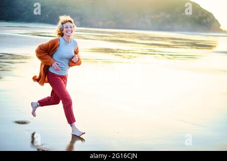 Jeune femme de race blanche, blonde et mûre, qui court à l'extérieur sur une plage par temps ensoleillé. Banque D'Images