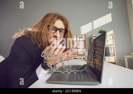 Travailleur de bureau malheureux assis à un bureau enchaîné à un ordinateur portable et incapable de rentrer à la maison Banque D'Images