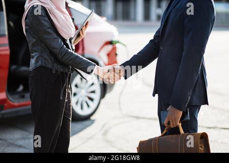 Un homme d'affaires africain qui se tremble avec une femme musulmane dans le hijab après un accord réussi. Charge de voiture électrique rouge en arrière-plan. Concept de personnes, vente et achat. Banque D'Images