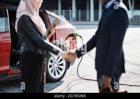 Flou avant-plan de la femme musulmane de vente de l'établissement de relations avec le client africain de l'extérieur. La voiture électrique rouge se recharge en arrière-plan. Homme d'affaires effectuant des achats coûteux. Banque D'Images