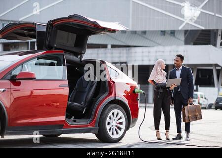 La voiture électrique rouge se recharge alors que la femme musulmane et l'homme africain parlent à proximité. Vendeur féminin dans hijab utilisant un ordinateur portable tout en faisant la démonstration de l'automobile moderne à un client masculin en costume. Banque D'Images