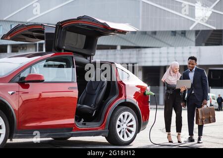 La voiture électrique rouge se recharge alors que la femme musulmane et l'homme africain parlent à proximité. Vendeur féminin dans hijab utilisant un ordinateur portable tout en faisant la démonstration de l'automobile moderne à un client masculin en costume. Banque D'Images