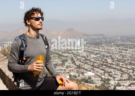 Jeune homme avec des lunettes de soleil tenant une bouteille d'eau et profitant de vues splendides sur la ville de Santiago au Chili le jour ensoleillé. Randonneur en train de faire une pause pendant la randonnée Banque D'Images