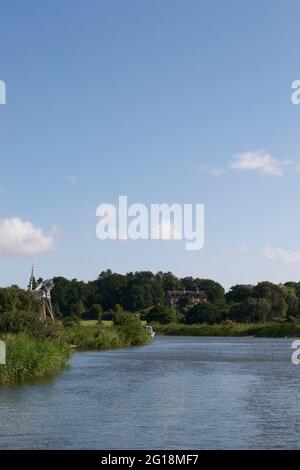 Un petit bateau à moteur qui se présente dans le virage dans une rivière tranquille et de campagne, le jour d'un été glorieux. Moulin à vent traditionnel vintage (pompe à vent) et un Banque D'Images