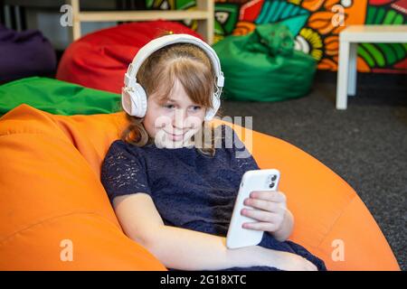 La petite fille utilise le téléphone et écoute de la musique avec un casque tout en étant assise sur un fauteuil à sac. Banque D'Images