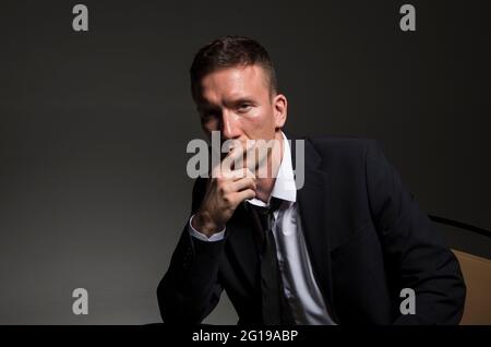 Portrait d'un homme confiant et pensif, dans un costume d'affaires, assis à pattes croisées sur un fond sombre. Banque D'Images